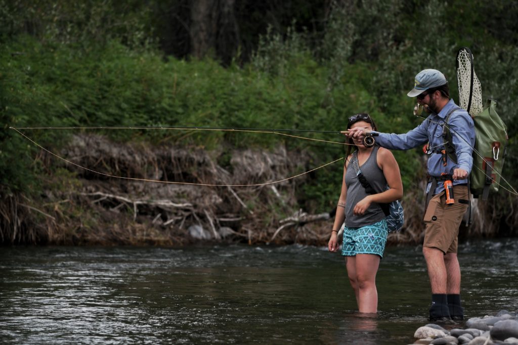A fly fishing guide shows a female client how to cast while standing in a river