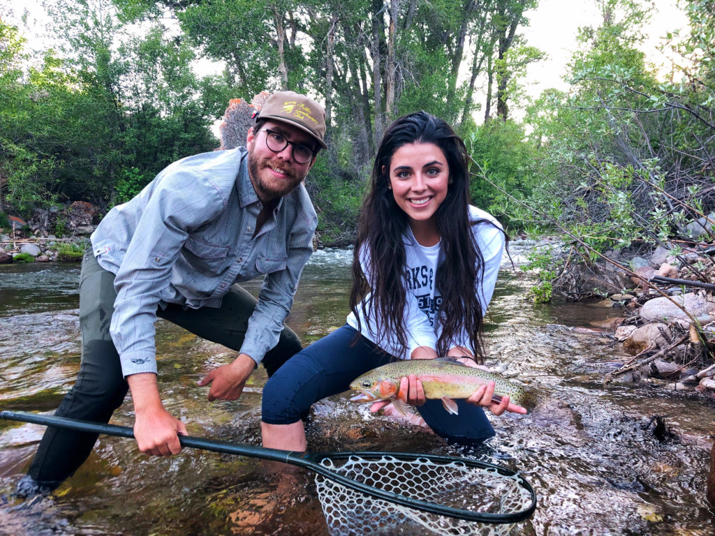 A fly fishing guide stands near a female client while she proudly shows a trout she just caught