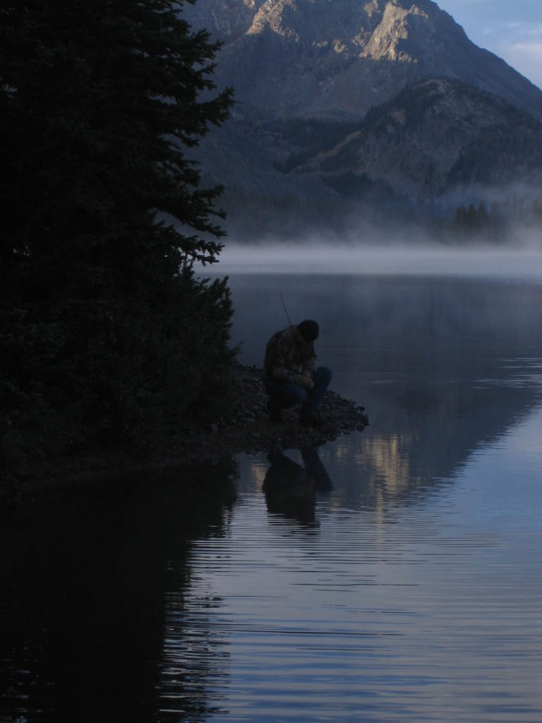 A man crouches by the side of an alpine lake at sunset on a horseback riding trip in Wyoming near CM Ranch