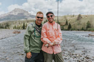 A fly fisherman proudly displays a trout he just caught while his friend stands by him and smiles